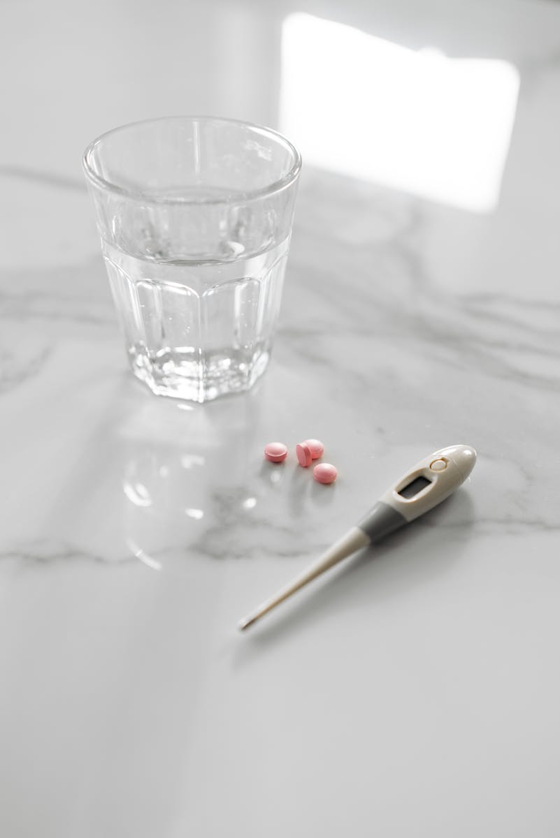 A glass of water with pills and a thermometer on a marble surface, ideal for health themes.